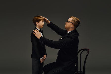 A father adjusts his son's bow tie, both dressed elegantly in tuxedos while posing in the studio.
