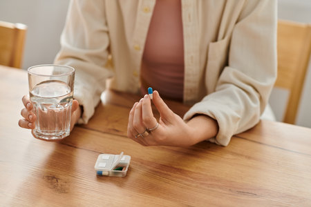 A woman holds a glass of water and a blue pill, sitting at a wooden table.のeditorial素材