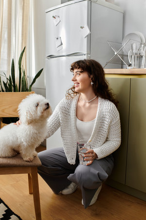 A young woman with curly hair smiles on the floor, holding a glass and petting her dog.のeditorial素材