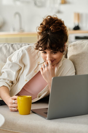 A young woman relaxes on a sofa, sipping coffee while engaging with her laptop during the afternoon.のeditorial素材