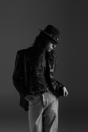 A handsome young man stands confidently, dressed in stylish attire, against a dark backdrop.