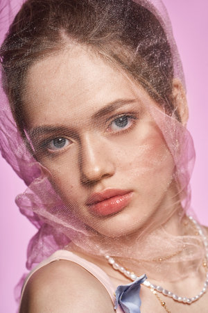 A stunning young woman with captivating features poses gracefully against a soft pink background.の写真素材