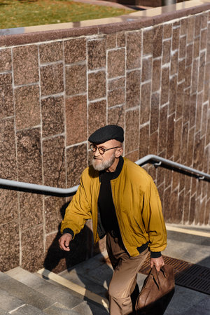 Stylish senior man walks confidently up the urban stairs in fashionable attire, showcasing contemporary trends against a cityscape backdropの写真素材