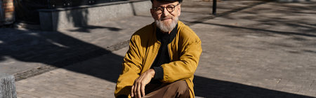 Charming senior man flaunts his stylish attire while enjoying a leisurely walk in the vibrant city landscape during a sunny dayの写真素材