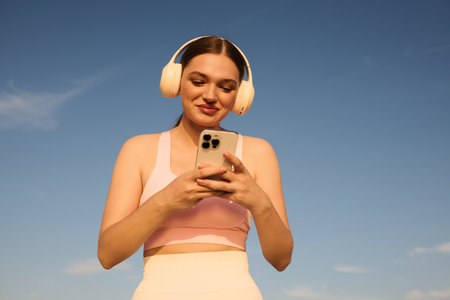 A smiling athletic woman in headphones checks her phone under a blue sky.の写真素材