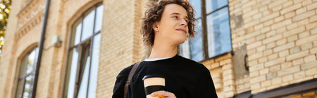 Young man with curly hair holds a coffee cup while walking past elegant brick buildings.のeditorial素材