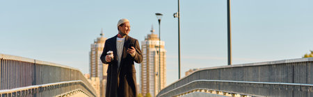Gentleman in trendy attire walks confidently on a bridge, sipping coffee while posing.