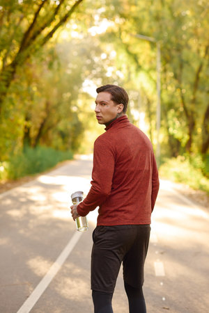 Tattooed man strolls a scenic road, sipping water amid colorful autumn trees.のeditorial素材