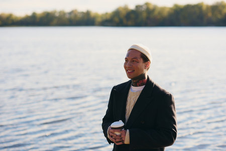 Handsome young man in a chic outfit smiles while holding coffee near tranquil water on a sunny day.