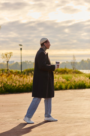 Handsome man with tattoos walks gracefully along a waterfront promenade, savoring coffee at dawn.の写真素材