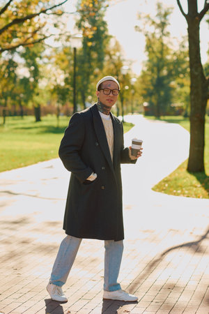 Handsome man dressed in modern attire walks through a park, sipping coffee under sunlight.