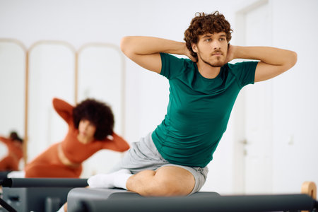 A man stretches on a reformer while a woman practices nearby in a bright studio.