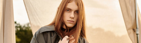 Young man with long hair shows off stylish attire against a sunset backdrop.