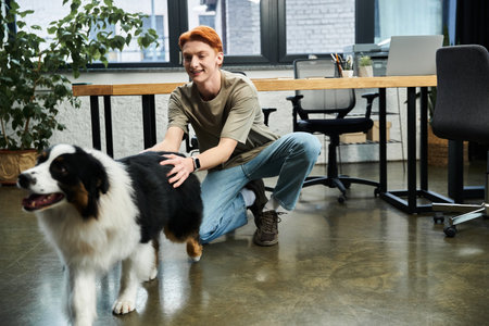 A young man with red hair kneels in an office, happily interacting with a friendly dog.のeditorial素材