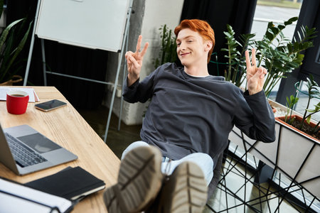 A young red haired man takes a break in a bright office, sitting casually and smiling at his desk.