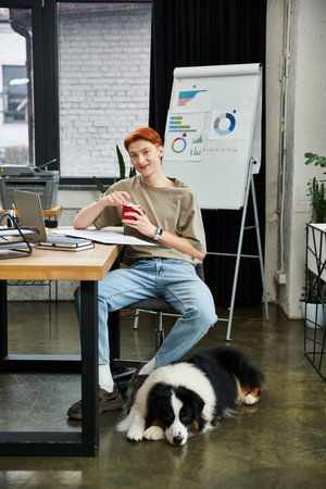 A young man with red hair relaxes at his desk in a lively office with his dog beside him.