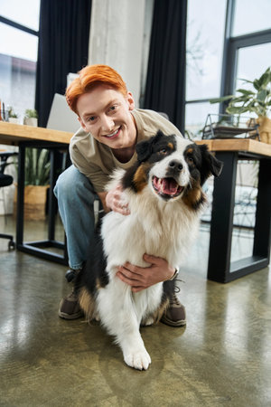 A dedicated young man with striking red hair smiles as he interacts with a friendly dog at work.