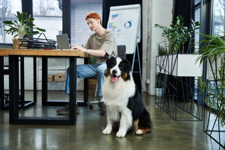 A dedicated young man with red hair focuses on his tasks at work while a cheerful dog sits nearby.のeditorial素材