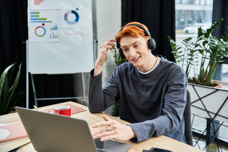 A handsome young man with ginger hair listens attentively while working on his laptop.