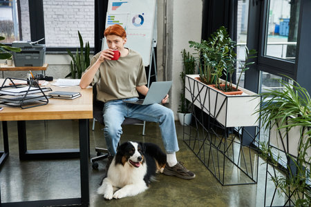 In a bright office, a red haired man takes a coffee break with a dog by his laptop.のeditorial素材