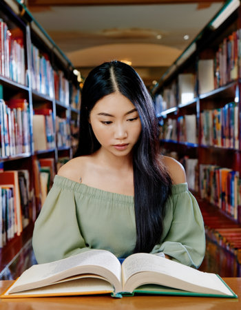 pretty asian student intensely reads a textbook in a quiet college library, surrounded by bookshelves.の素材