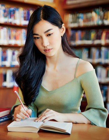 A young asian student diligently takes notes while studying in a quiet library setting filled with books.の素材