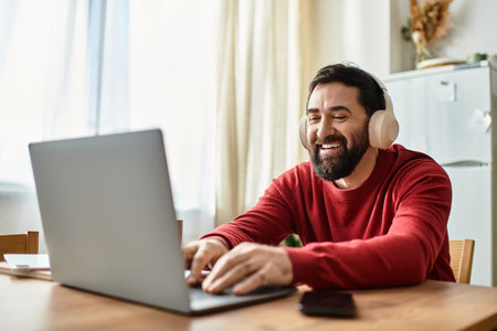 Cheerful older man listens to music with headphones while typing on his laptop at homeのeditorial素材
