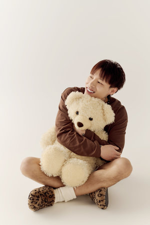 Handsome young asian man smiles brightly while hugging a large teddy bear in a cozy setting.