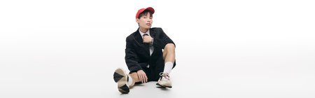 A young man showcases a relaxed demeanor, seated comfortably in a trendy outfit with a cap.