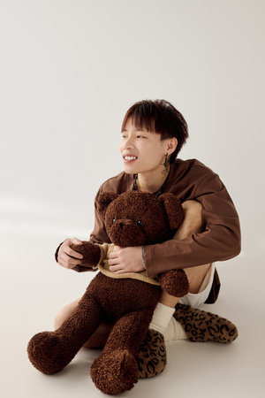 A joyful young Asian man holds a large teddy bear in a warm, minimalistic setting.の写真素材