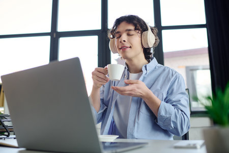 In a bright office, a young man savors a warm beverage while listening to music with headphones.