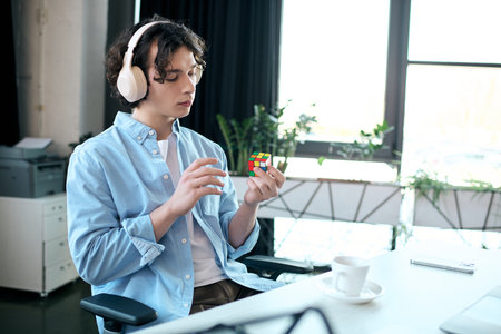 A focused young man wearing headphones works with a Rubiks cube in a stylish office setting.