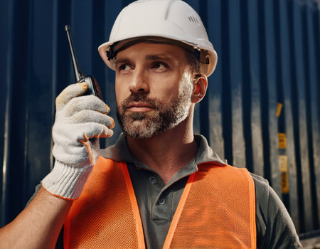 Construction worker in safety gear uses a radio to coordinate tasks amid ongoing operations.の素材