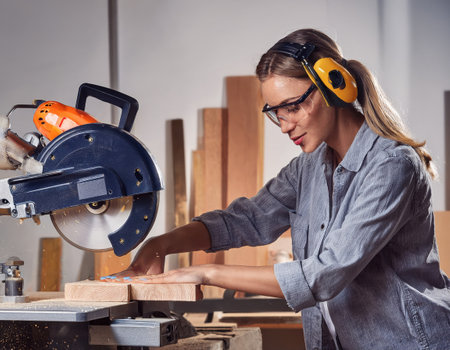 A skilled female worker uses a power saw to shape wood in a busy construction workshop.の素材