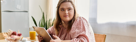 Young woman with Down syndrome sits at a table, enjoying juice and using a tablet.のeditorial素材