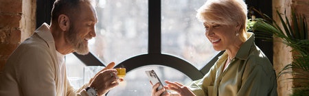 A middle aged couple shares laughter and stories over drinks while looking at a smartphone, banner
