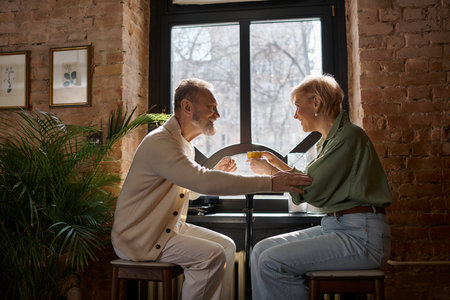 A middle-aged couple shares laughter and drinks, enjoying their intimate time by the window.のeditorial素材