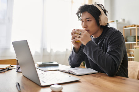 A handsome young asian man is focused on his studies, using a laptop while enjoying a warm drink.