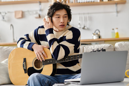 Handsome young Asian man focuses on learning acoustic guitar while seated comfortably indoors.のeditorial素材
