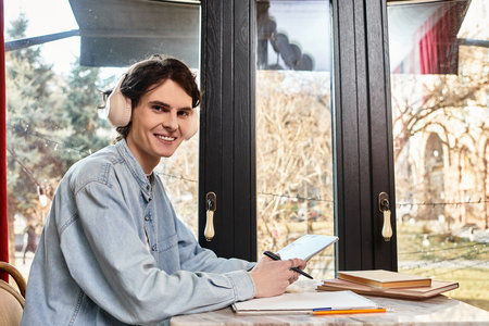 Cheerful young man with headphones sits at a table, studying and enjoying a calm cafe atmosphere.