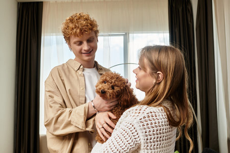 A young couple smiles at each other, holding their adorable dog in a hotel room.