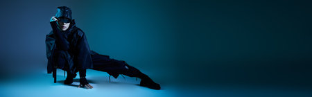 A handsome young man in dark attire poses confidently on a chair under blue lighting.の写真素材