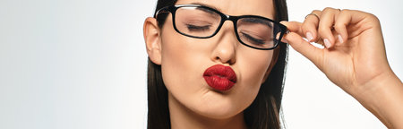 A young woman with brown hair and red lips playfully adjusts her glasses on a grey backdrop, bannerのeditorial素材