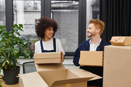 Two people joyfully sort moving boxes in a modern office with nearby plants.