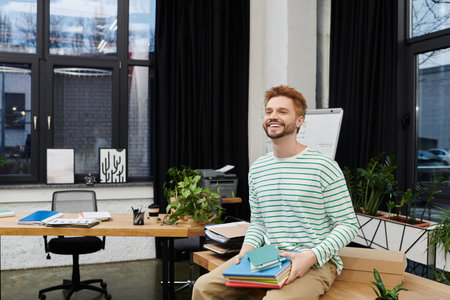 A cheerful man arranges a stylish office with a stack of colorful books.のeditorial素材