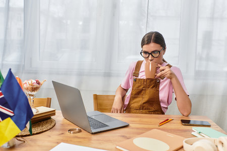 A young beautiful woman sips coffee at her desk while focused on her laptop and notes.