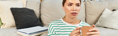 A young beautiful woman sits comfortably on her couch, holding a warm drink while deep in thought.のeditorial素材