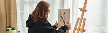 A young woman with Down syndrome sets up art on an easel in her cozy, sunlit home.のeditorial素材