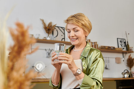 Beautiful senior woman smiles as she admires a glass of water in a warm and inviting space.のeditorial素材