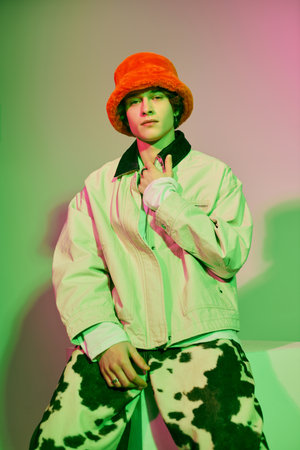 Young man poses confidently in trendy attire, lit by colorful lights in a creative backdrop.のeditorial素材